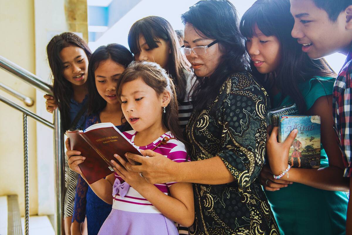 Child from Indonesia reading her Bible with family surrounding her.